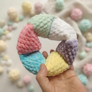 Pode incluir: Um anel circular de crochê feito de fio macio e felpudo em um padrão de arco-íris segmentado. O anel apresenta seções alternadas de verde menta pastel, branco, lavanda, amarelo pálido, azul brilhante e rosa suave, com uma aparência texturizada.