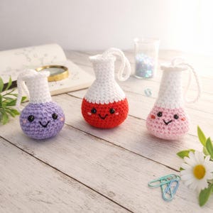 May include: Three crocheted flask ornaments in purple, red, and pink, each with a white top and a smiling face. The flasks have a loop for hanging. The background includes a notebook, a magnifying glass, and a daisy.