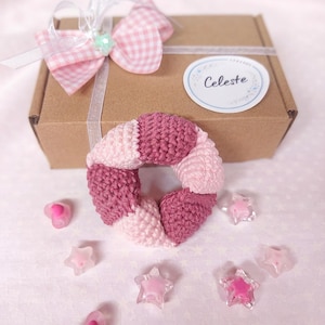 Puede incluir: Un anillo de ganchillo con segmentos alternos de hilo rosa claro y rosa oscuro, colocado frente a una caja de regalo de cartón marrón atada con una cinta blanca y un lazo de cuadros rosa. La caja tiene una etiqueta circular con el texto Celeste.