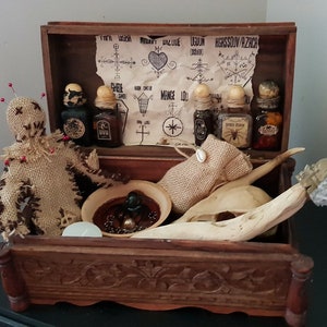 May include: A dark brown wooden box with carved detailing, containing occult items. Inside are a burlap doll with pins, small bottles, a skull, a seashell, and a scroll with symbols. The box is open.
