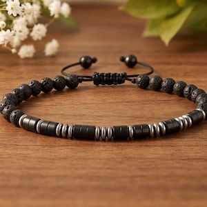 Könnte beinhalten: Ein Armband mit schwarzen, porösen Lavasteinperlen und abwechselnden zylindrischen schwarzen und silberfarbenen Metallabstandshaltern. Das Stück verfügt über eine schwarze Kordel mit verstellbarem Schiebeknotenverschluss und zwei kleinen schwarzen Perlen an den Enden.