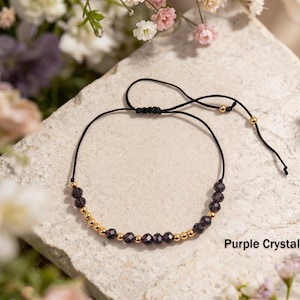 May include: A delicate adjustable bracelet featuring a black cord with a sliding knot closure, accented by alternating small gold-toned metallic beads and faceted dark purple crystal beads. The text Purple Crystal is visible.