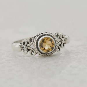 Puede incluir: Anillo de plata con una piedra preciosa redonda de color amarillo dorado en el centro. La piedra preciosa está rodeada por un borde texturizado en forma de cuerda. El anillo presenta filigranas decorativas en espiral a ambos lados de la piedra.