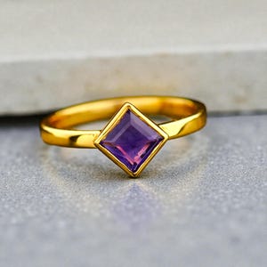 May include: A gold ring with a square, purple gemstone. The ring's band is a polished gold color, and the gemstone is set in a square bezel. The ring is displayed on a gray surface.