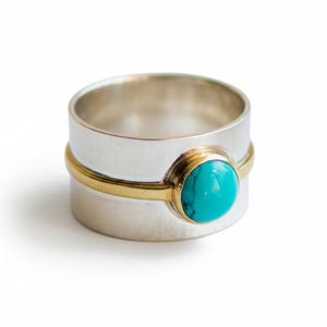 Puede incluir: Un anillo ancho de plata con una banda dorada y una piedra turquesa. El anillo tiene una banda gruesa y un acabado pulido. La piedra turquesa es de forma ovalada y está engastada en un bisel dorado. Fondo blanco.