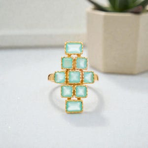 Pode incluir: Um anel dourado com um conjunto de pedras preciosas retangulares azul claro. As pedras estão dispostas em um padrão semelhante a uma cruz, com uma fileira central e pedras adicionais acima e abaixo. Em um fundo macio e neutro.