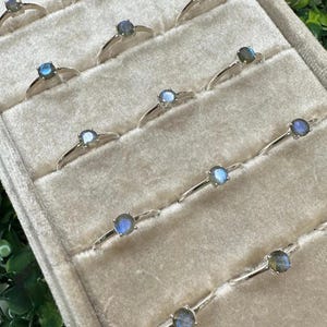 May include: A collection of silver rings, each featuring a small, round, blue gemstone. The rings are displayed in a light-colored, velvet-lined jewelry tray. The gemstones have a subtle, iridescent quality, reflecting various shades of blue and green.
