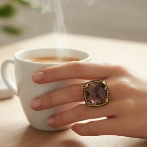 以下が含まれることがあります： 金色のフレームと黒いアクセントが付いた、大きな正方形カットのスモーキークォーツリングをはめた手。手は白いコーヒーカップを持っており、飲み物から湯気が上がっています。リングが焦点です。
