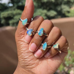 May include: Five silver rings each feature a unique, raw-cut opal gemstone. The opals display a range of colors, including blue, green, and yellow, set against a silver band. The rings are displayed on a hand.