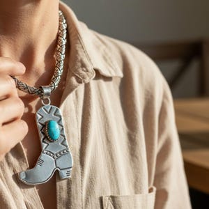 May include: Silver-toned pendant necklace featuring a cowboy boot design with a turquoise stone. The boot pendant is detailed with etched patterns and hangs from a thick, braided chain. The necklace is worn against a beige shirt.