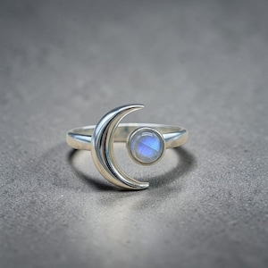 Puede incluir: Un anillo de plata con un diseño de media luna y una piedra preciosa azul redonda. El anillo tiene un acabado pulido y está sobre un fondo gris. La piedra preciosa tiene una sutil cualidad iridiscente.