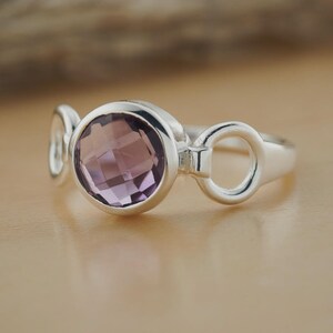 Op de afbeelding: Zilveren ring met een grote, ronde, gefacetteerde amethist edelsteen. De ring heeft een uniek ontwerp met een cirkelvormig element aan weerszijden van de edelsteen, wat een moderne en elegante uitstraling creëert. De amethist is lichtpaars.