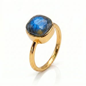 Peut inclure: Une bague dorée ornée d'une pierre de labradorite à facettes de forme carrée avec des reflets bleus irisés. La pierre est sertie dans une monture en métal doré lisse et poli, fixée sur un anneau fin, simple et arrondi en métal doré.