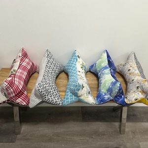 May include: Five decorative pillows in a fishbone shape rest on a wooden bench with metal legs. The pillows feature various fabric patterns, including red plaid, gray swirls, blue floral, and a watercolor design. The pillows are a decorative home accent.