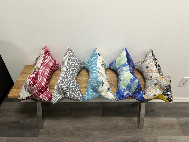 May include: Five decorative pillows in a fishbone shape rest on a wooden bench with metal legs. The pillows feature various fabric patterns, including red plaid, gray swirls, blue floral, and a watercolor design. The pillows are a decorative home accent.