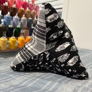 May include: A triangular-shaped pillow with a patchwork design. One section features a black and white plaid pattern, another has white feathers on a black background, and the third has musical notes on a black background. The pillow is resting on a blue and white striped surface.