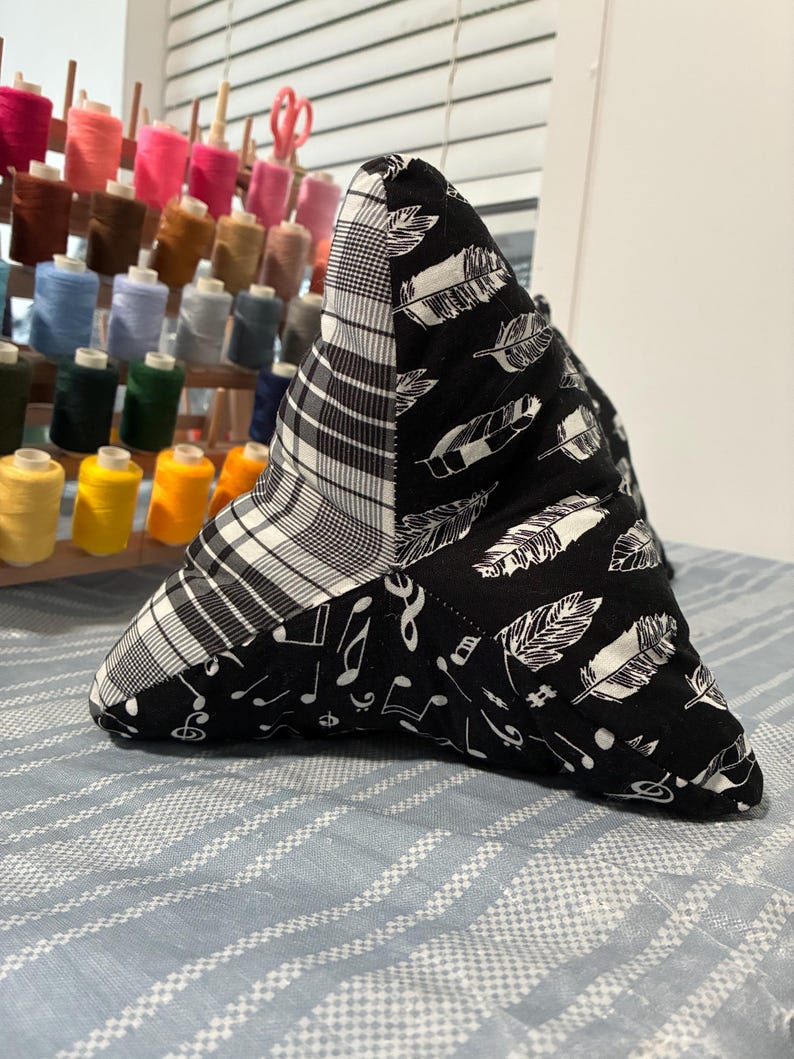 May include: A triangular-shaped pillow with a patchwork design. One section features a black and white plaid pattern, another has white feathers on a black background, and the third has musical notes on a black background. The pillow is resting on a blue and white striped surface.