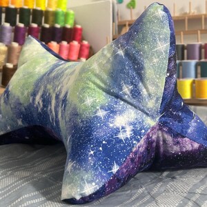 May include: A star-shaped reading pillow with a galaxy print in shades of blue, purple, and green. The pillow is made of soft fabric and features white star accents. It is designed to provide support while reading.