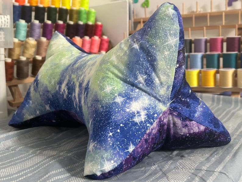May include: A star-shaped reading pillow with a galaxy print in shades of blue, purple, and green. The pillow is made of soft fabric and features white star accents. It is designed to provide support while reading.