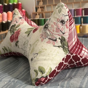 May include: A decorative, butterfly-shaped pillow with floral and butterfly patterns. The pillow features a cream-colored background with red and green floral accents. The edges are trimmed with a red and white plaid and a dark red quatrefoil pattern.