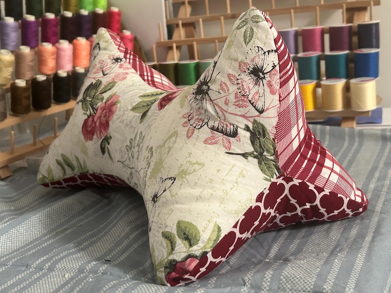 May include: A decorative, butterfly-shaped pillow with floral and butterfly patterns. The pillow features a cream-colored background with red and green floral accents. The edges are trimmed with a red and white plaid and a dark red quatrefoil pattern.
