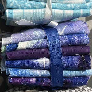 May include: Stacks of fabric squares in various colors and patterns, including yellow, blue, purple, gray, and red. The fabric bundles are tied with ribbons in white, blue, and gray. Ideal for quilting or crafting projects.
