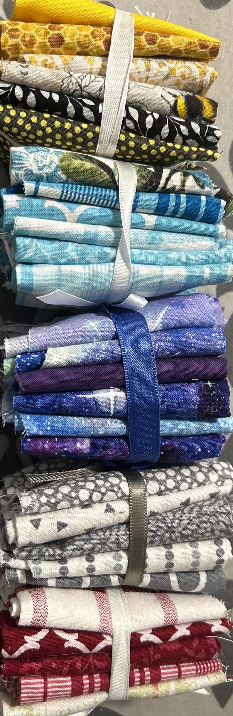 May include: Stacks of fabric squares in various colors and patterns, including yellow, blue, purple, gray, and red. The fabric bundles are tied with ribbons in white, blue, and gray. Ideal for quilting or crafting projects.