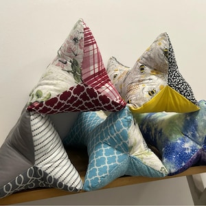 May include: A collection of handmade triangular book pillows in various patterns. Designs include floral, plaid, bee, geometric, and galaxy prints. The pillows are arranged on a wooden surface.