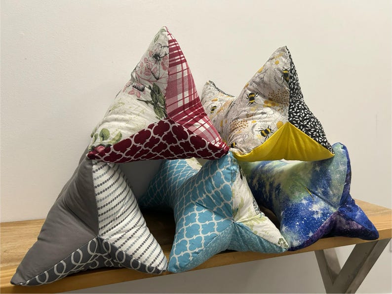 May include: A collection of handmade triangular book pillows in various patterns. Designs include floral, plaid, bee, geometric, and galaxy prints. The pillows are arranged on a wooden surface.