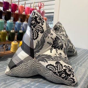 May include: Two star-shaped decorative pillows. One pillow features a black and white floral pattern, a gray and white plaid, and a gray chevron pattern. The other pillow has a similar floral pattern. The pillows are on a blue and white striped surface.