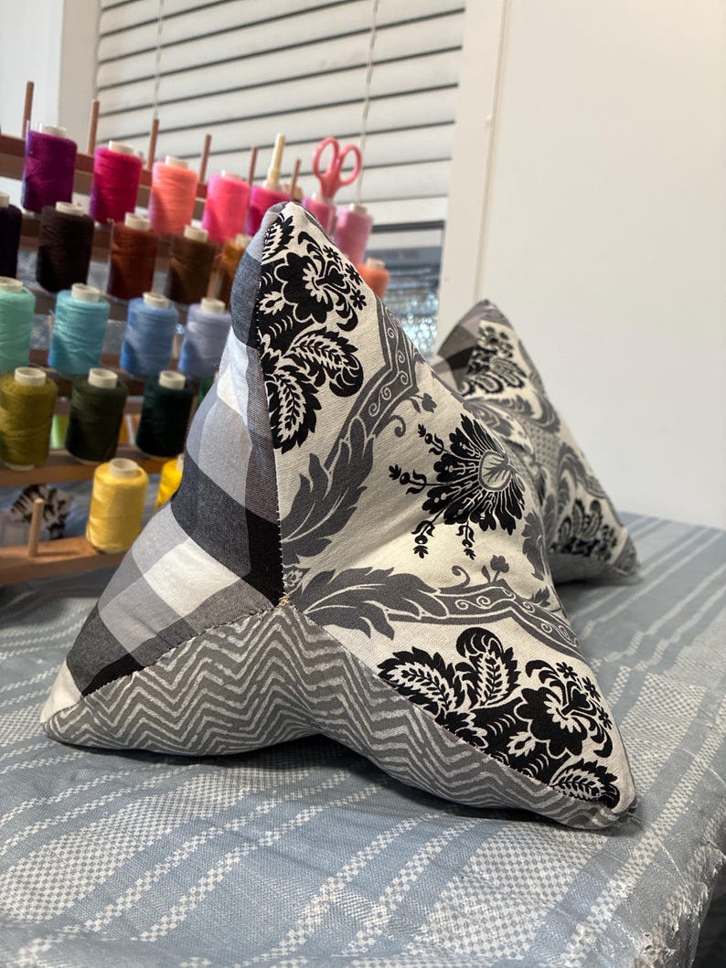 May include: Two star-shaped decorative pillows. One pillow features a black and white floral pattern, a gray and white plaid, and a gray chevron pattern. The other pillow has a similar floral pattern. The pillows are on a blue and white striped surface.