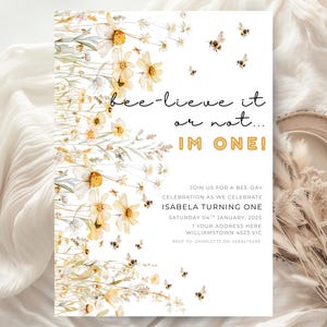 Peut inclure: Une invitation blanche avec des illustrations florales et d'abeilles. Le texte dit "bee-lieve it or not... I'M ONE!" et invite les invités à une fête d'anniversaire. La conception comprend des marguerites à l'aquarelle et des abeilles volantes.