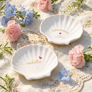 May include: Two white ceramic shell-shaped trinket dishes. One dish features the gold text EMILY & MIKE, a red heart, and Engaged September 5, 2020. The other dish features the gold name isabella in script with a small red heart above it.