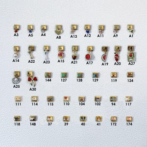 May include: A collection of gold-toned charms with various designs, including a star, heart, bow, and fruit. Each charm has a small clasp for attachment. The charms are arranged on a white surface, with each charm labeled with a letter and number.