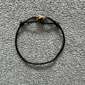 May include: A black cord bracelet with a gold-colored, intertwined knot design at the center. The bracelet is set against a textured, gray background, highlighting the contrast between the dark cord and the metallic accent.