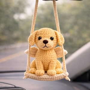 May include: A crocheted yellow puppy car charm with small wings, suspended from a beige rope swing. The puppy has black eyes and a brown nose. The charm is hanging from a car's rearview mirror.