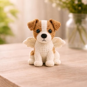 May include: A crocheted dog figurine with brown and white markings, large black eyes, and small white wings. The handmade dog is sitting on a wooden surface. A vase of white flowers is in the background.