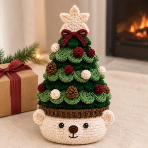 Puede incluir: Una decoración de árbol de Navidad de ganchillo con un diseño de niveles en verde oscuro, adornado con pequeñas esferas rojas y blancas y piñas marrones. El árbol está rematado con una estrella color crema y un lazo rojo sobre una base de oso.