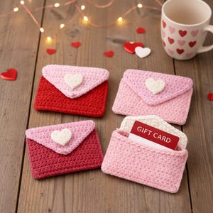 Può includere: Quattro portacarte regalo fatti a mano all'uncinetto, a forma di busta, nei toni del rosso e del rosa, ciascuno con un cuore bianco. Un portacarte è aperto, mostrando una carta regalo. Una tazza con disegni a cuore e decorazioni a forma di cuore completano la scena.