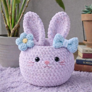 Peut inclure: Un panier lapin en crochet fait à la main, de couleur lavande, avec des oreilles dressées, un nœud bleu clair et une fleur bleue. Le panier a un nez rose, des yeux noirs et des moustaches roses. Le panier est sur une surface lavande.