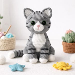 May include: A handmade crochet cat in shades of gray and white, with black stripes and green eyes. The cat is sitting upright, with a white chest and paws. A blue starfish and yellow sea creature are in the foreground.