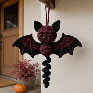 Pode incluir: Um ornamento de morcego em crochê com cabeça e corpo em bordô escuro, com asas e orelhas pretas. O morcego possui duas pequenas presas brancas, olhos de segurança pretos e uma longa cauda preta em espiral. Um laço bordô está preso no topo para pendurar.