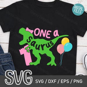 May include: A black t-shirt featuring a green dinosaur graphic with a pink bow on its head. The text reads ONE a SAURUS with a large pink number 1. Three balloons in yellow, pink, and blue are attached to the dinosaur tail. A digital files label is visible.