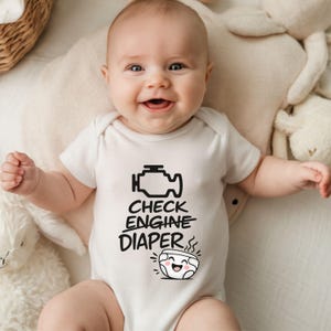 May include: A baby in a white onesie with the text "CHECK ENGINE DIAPER" and a graphic of an engine and a smiling diaper. The baby is smiling. The background includes a cream-colored surface and soft toys.