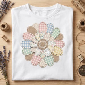 Könnte beinhalten: Weißes T-Shirt mit einem großen Blumenmuster. Die Blume besteht aus Stoffflicken in Pastelltönen und Vichy-Mustern, mit einer Mitte aus Jute. Das Design ist auf das T-Shirt genäht und erzeugt einen strukturierten Effekt.