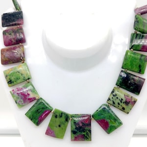 Può includere: Una collana con perline rettangolari verdi, rosa e nere. Le perline sono collegate con connettori color oro, creando un accessorio unico e colorato. La collana è esposta su un manichino bianco.