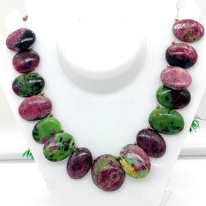 May include: A necklace with large, oval beads in deep red, green, and black. The beads are connected with gold-coloured links. The necklace is displayed on a white mannequin, showcasing the vibrant colours and unique design.