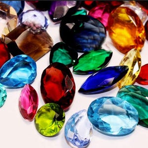 Op de afbeelding: Een verzameling gefacetteerde edelstenen in verschillende vormen en kleuren, waaronder rood, blauw, groen, geel en paars. De edelstenen zijn verspreid, wat hun schittering en helderheid benadrukt. Ideaal voor het maken van sieraden.