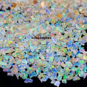 May include: A collection of small, irregular opal gemstone chips featuring a translucent white base with vibrant flashes of blue, green, and pink iridescence. The text RAINBOWGEMS is printed in black in the centre of the scattered gemstone pile.