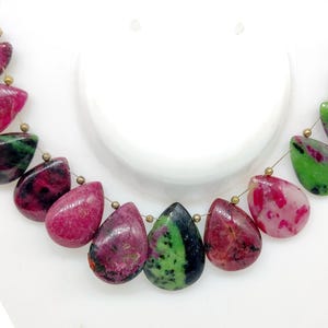 Op de afbeelding: Een ketting met druppelvormige kralen in rood, groen en zwart. De kralen zijn geregen met goudkleurig draad, wat een uniek en opvallend sieraad creëert. De ketting wordt gepresenteerd op een witte buste.